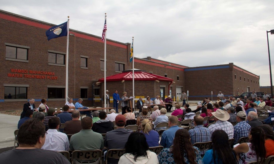 Photo of a large audience of people outdoors with a brick building and flags on flag poles