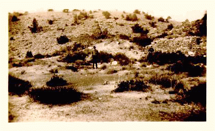 A man standing on the site of an old copper smelter. Hill side is a barren area dotted sparcely with sagebrush. Sepia tone photo