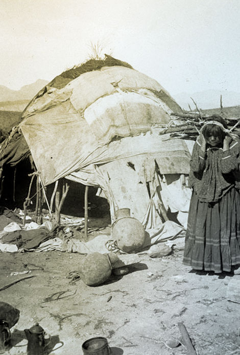 Apache workers often brought their families to live with them. They lived in a segregated area and maintained their traditional lifestyle.  Here an Apache woman brings firewood to her wickiup, a structure of bent poles covered with brush that is also covered with blankets, tarps, and hides.  Note the use of large ceramic storage jars and metal pots and pans, 1911. (Reclamation photograph)