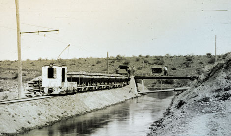 An electric engine is hauling cars filled with clay for use in the construction of Roosevelt Dam.  The tracks are adjacent to the power canal that was built to divert water from the Salt River and carry it to a penstock near the dam site where it fell 220 feet to power a water turbine.  The penstock and turbine were temporarily located in small cave south of and below the dam site. Electricity generated by the turbine provided power for this engine and a host of other tools and appliances at the dam site and the nearby town of Roosevelt, 1905. (Restoration photograph)
