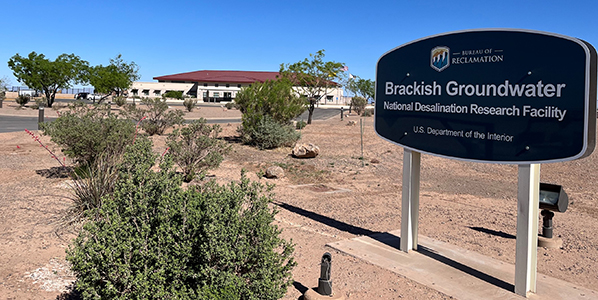 A photo of the sign at the Brackish Groundwater National Desalination Research Facility with the facility in the facility in the background. The sign is surrounded by desert landscaping.