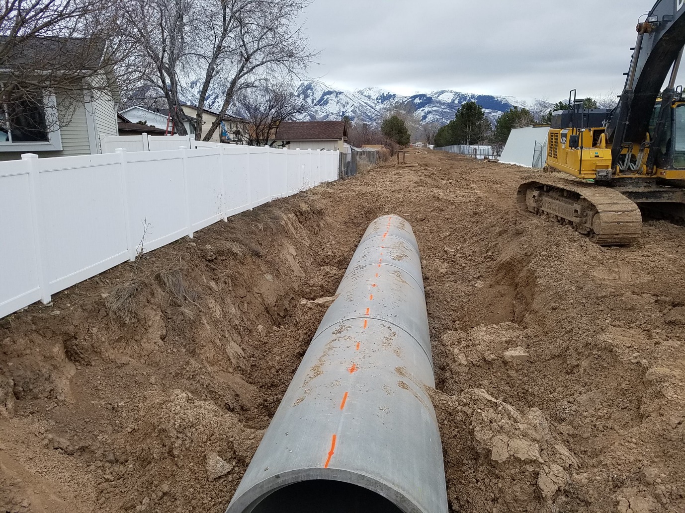 A pipe laying in dirt with a white fence on the left and an excavator on the right.