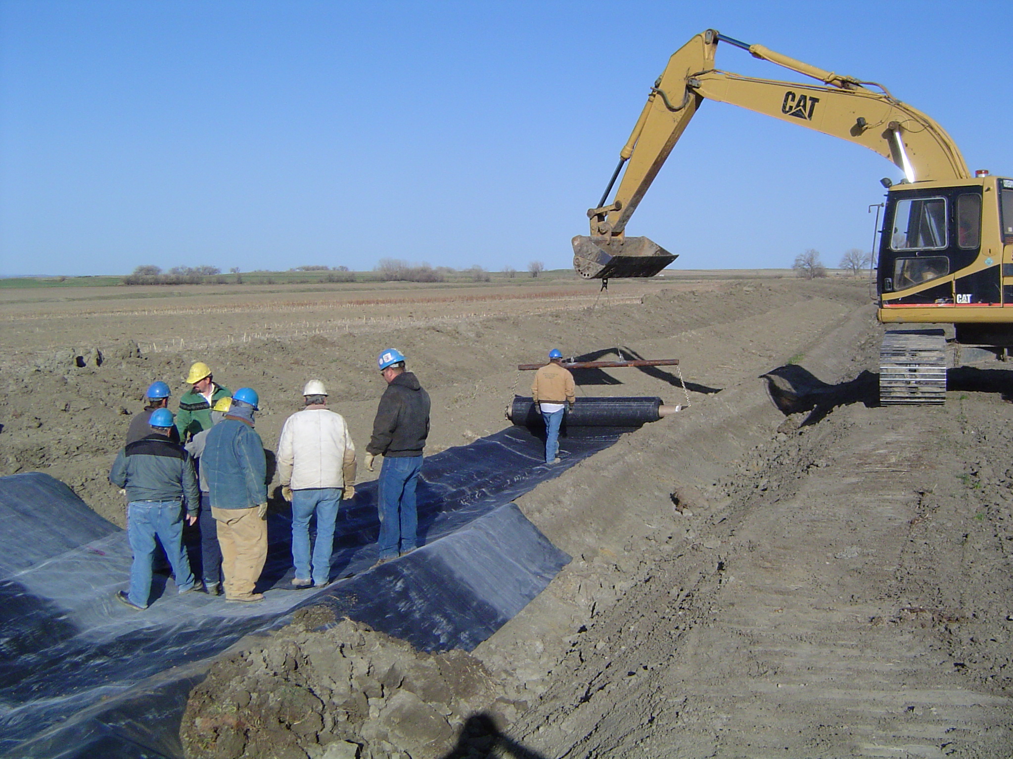 An excavator is helping people roll out a lining in a small canal in a field.