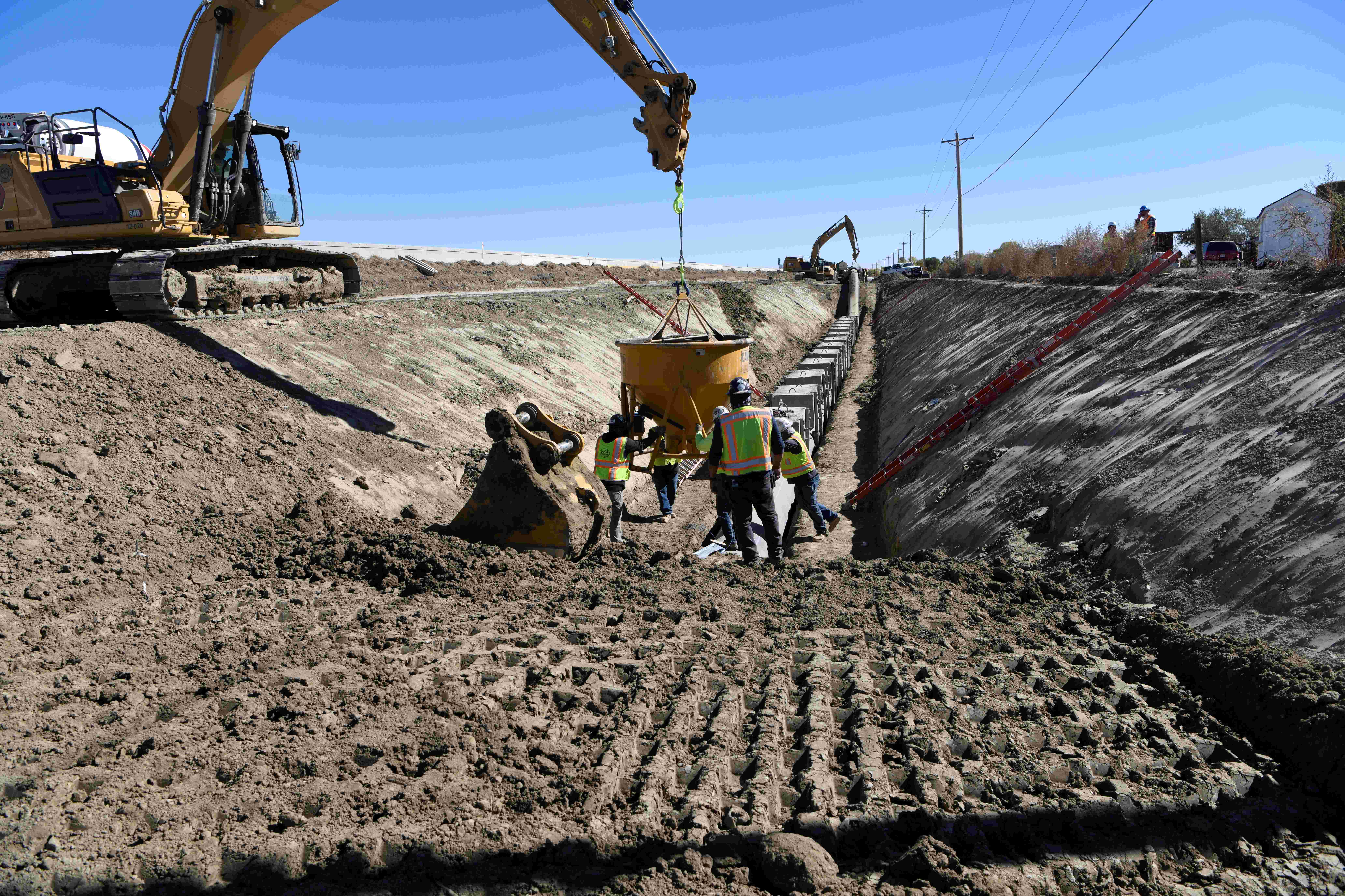 People working in a trench installing pipe. An excavator is lowering a manifold down into the trench.