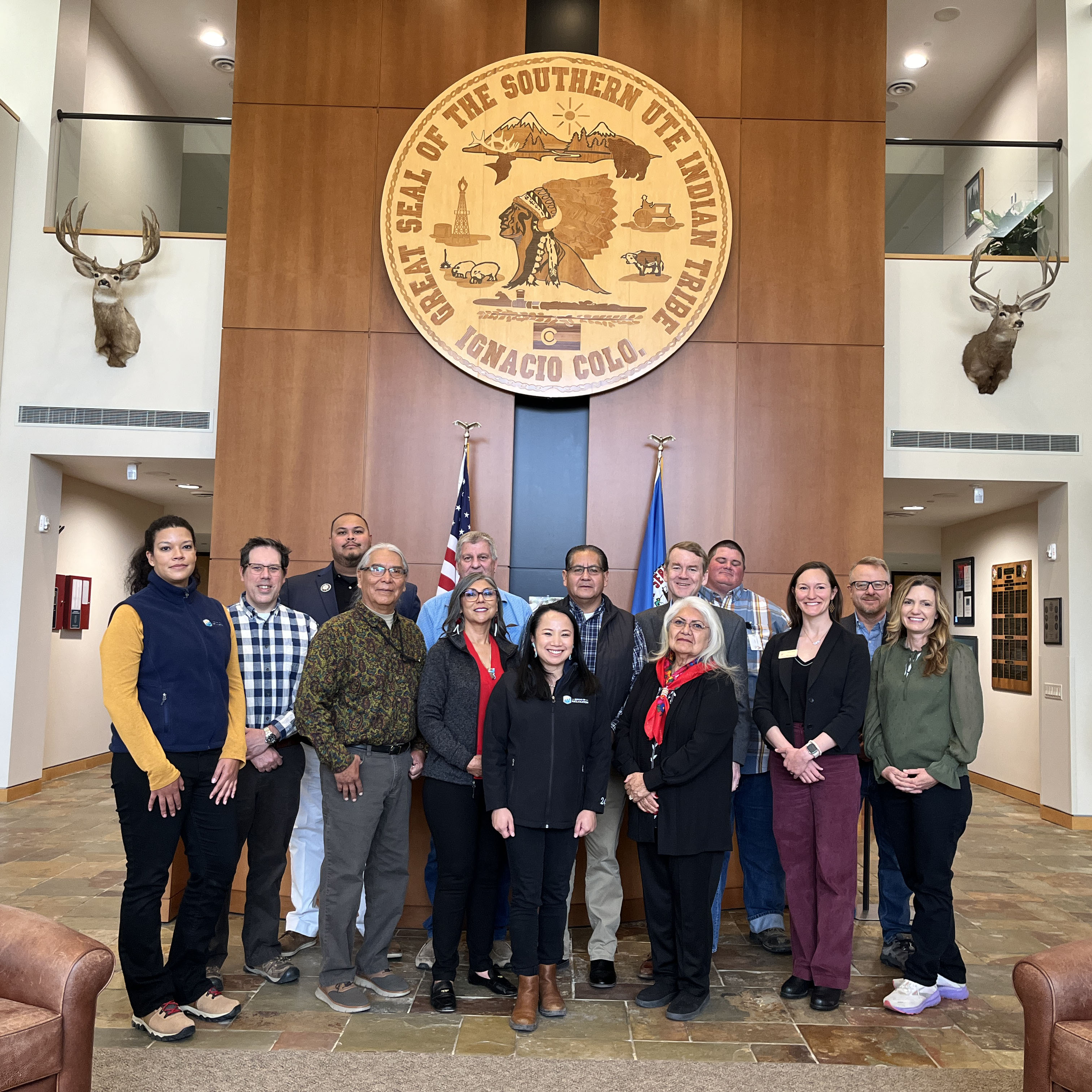 A group of people standing in a building in front of a big wall. On the wall is a large oval that says the Great Seal of the Southern Ute Indian Tribe in Ignacio, Colorado.
