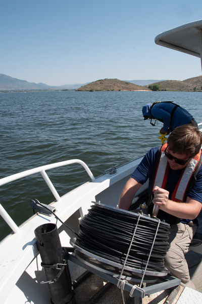 In the foreground Mitch Tree has his hand on the handle of the lowering wheel for the Hydrolab probe and behind him along the side rail of the boat Alex Walker is leaning over the side attempting to see the Secchi wheel that has been lowered into the reservoir.
