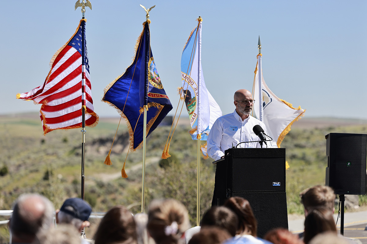 Man in white shirt speaking at outdoor podium with American and state flags behind him against a desert landscape.