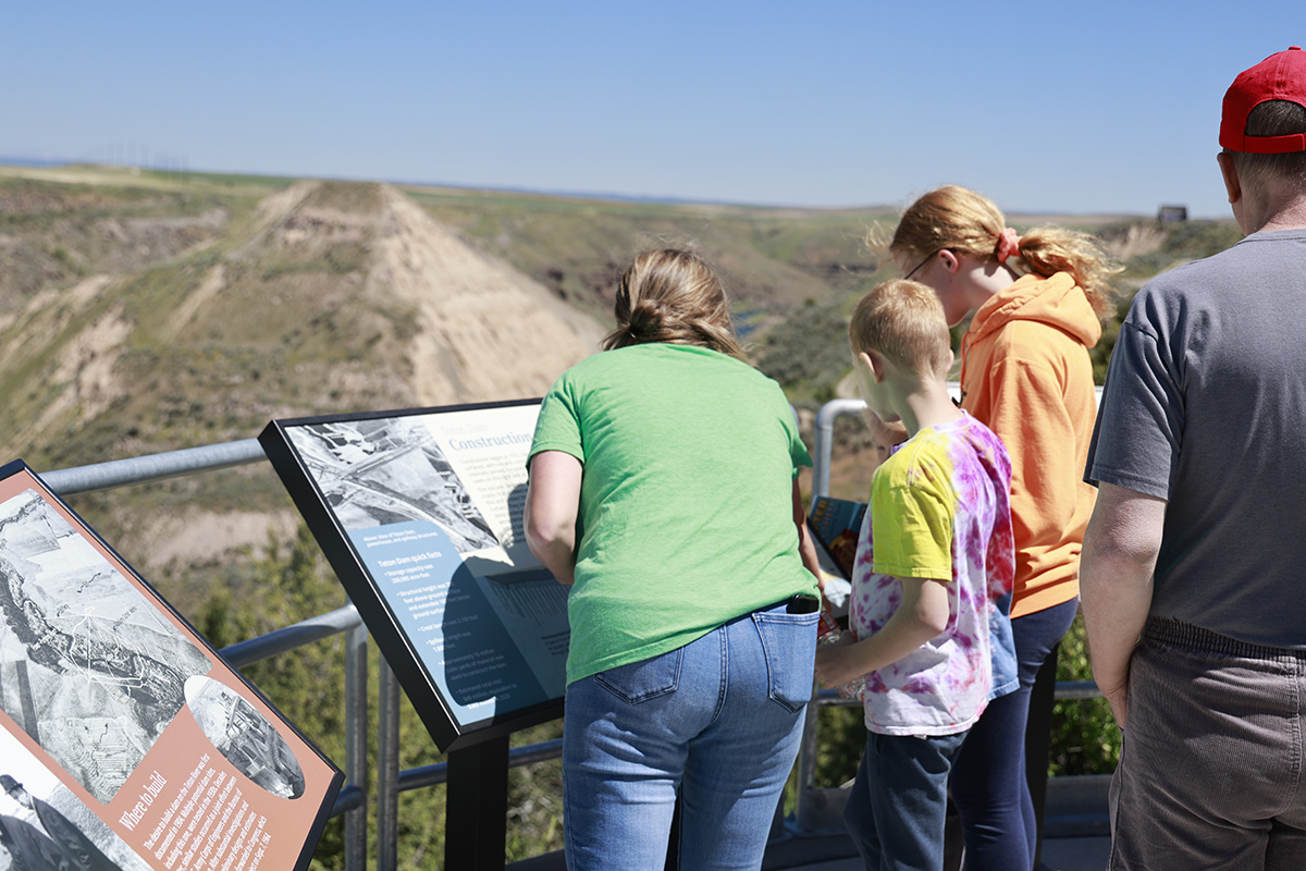Group of people reading informational signs at a scenic overlook with hills in the background under clear blue sky.