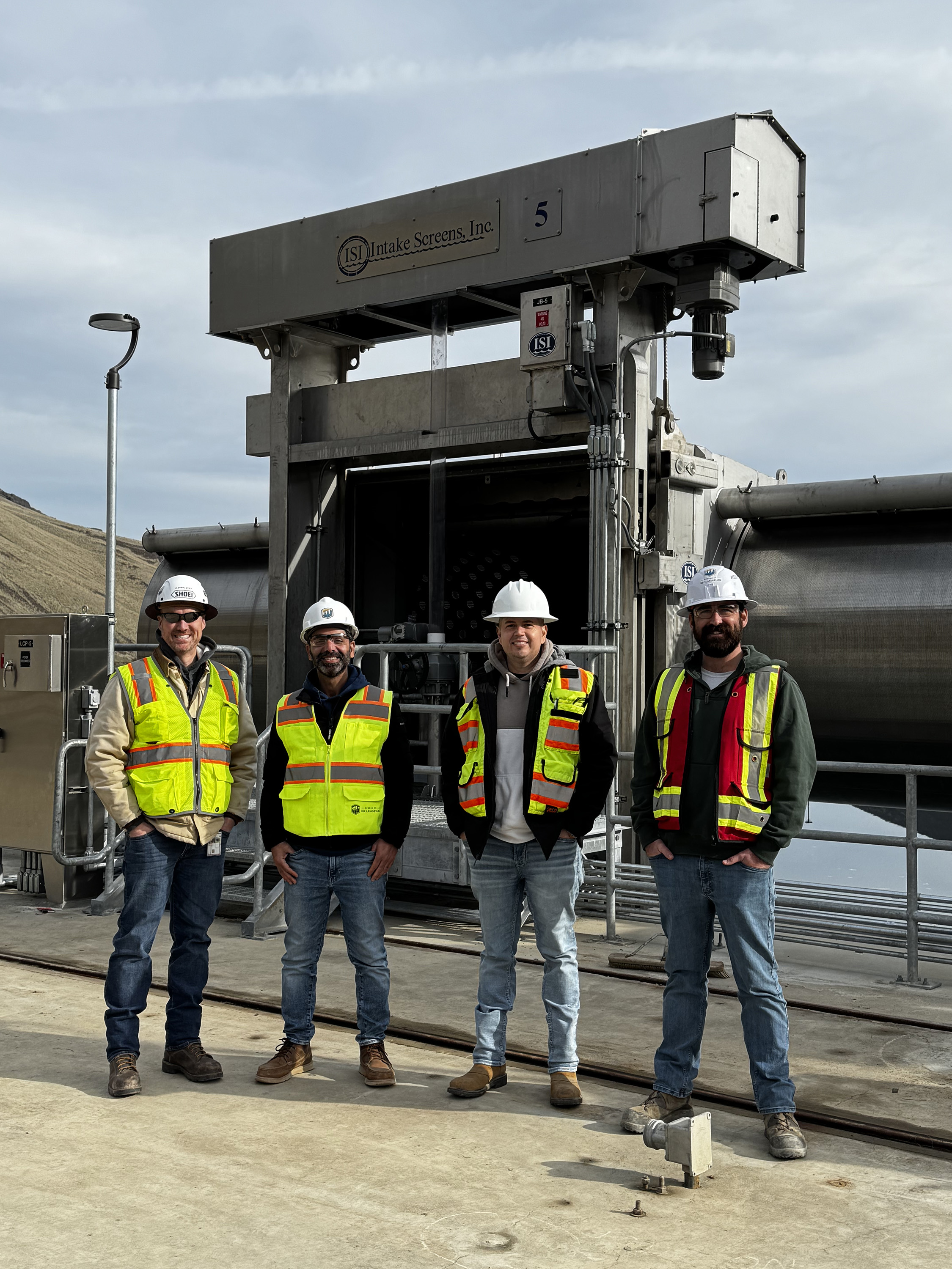 Four individuals wearing safety vests and helmets stand in front of large industrial machinery outdoors.