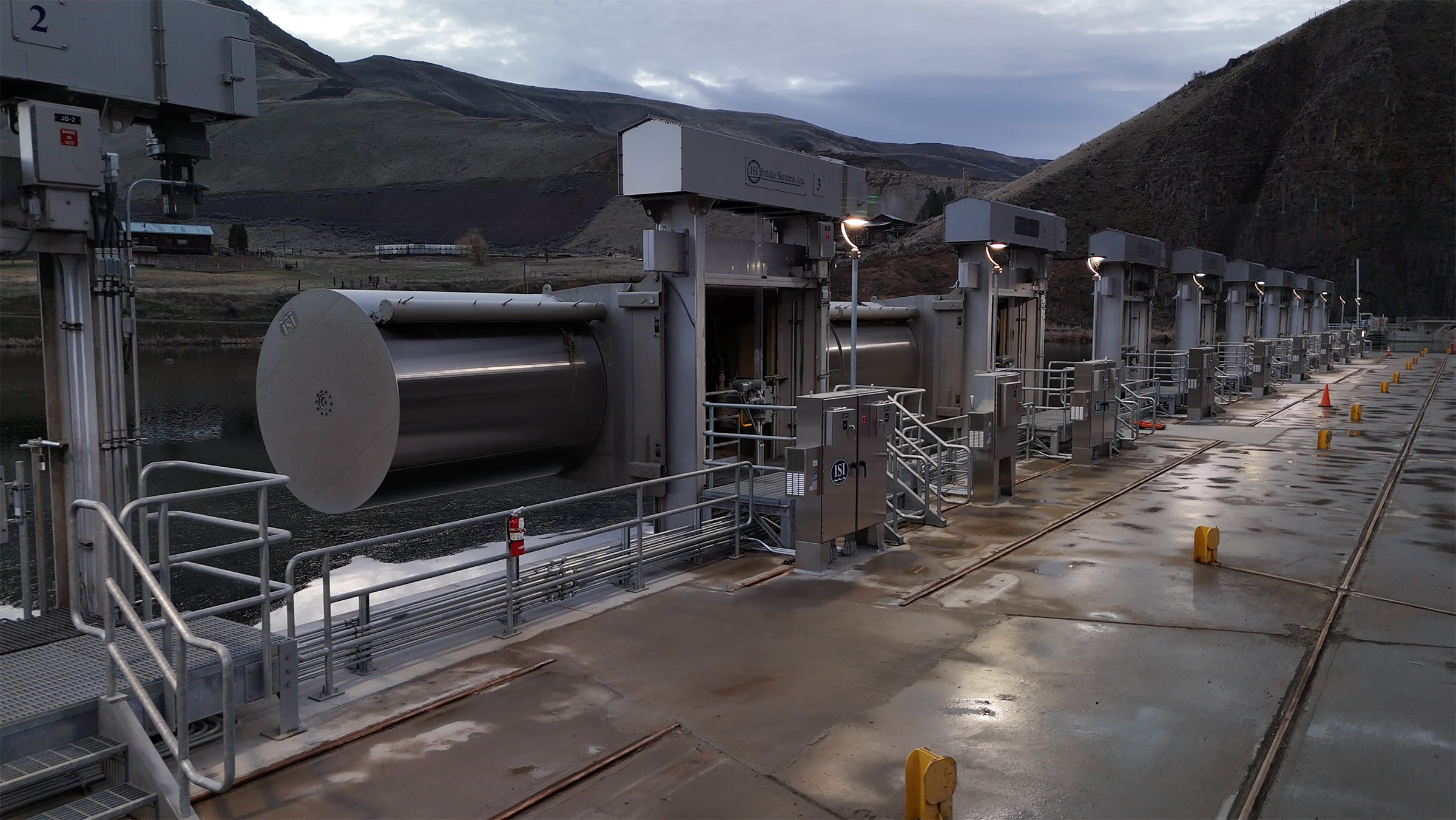 Long row of industrial floodgates with metal railings and control booths along a water channel in a mountainous area under cloudy sky.