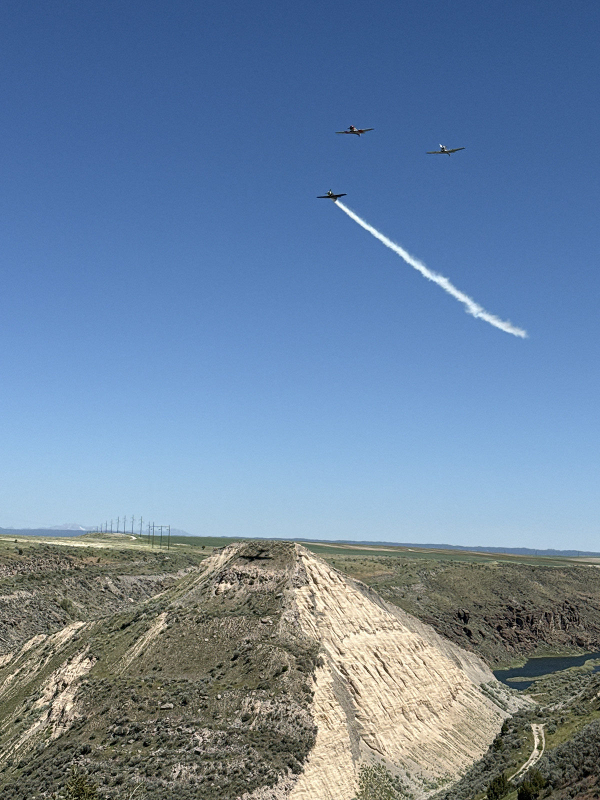 Three small airplanes flying in formation over a rugged canyon landscape with one plane trailing white smoke against a clear blue sky.