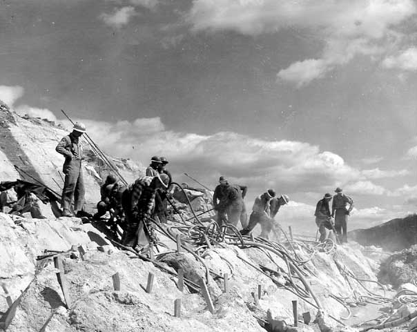 Jackhammer crews operating high on canyon wall preparing for construction of parking area retaining wall.