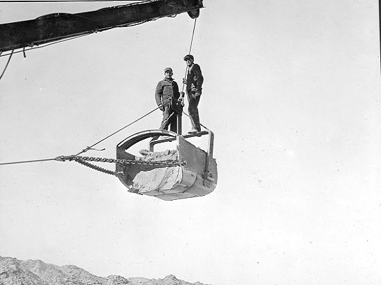Two men standing on a suspended seam engine bucket full of dirt.