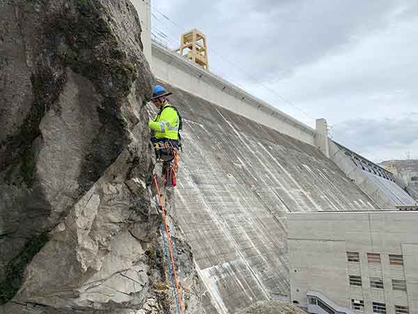 Reclamation employee Jerald “Denny” Womack is suspended halfway down the cliff face doing rock scaling and vegetation removal above the parking lot between the Right and Third Powerplant. Background is Grand Coulee downstream side of dam and spillway.