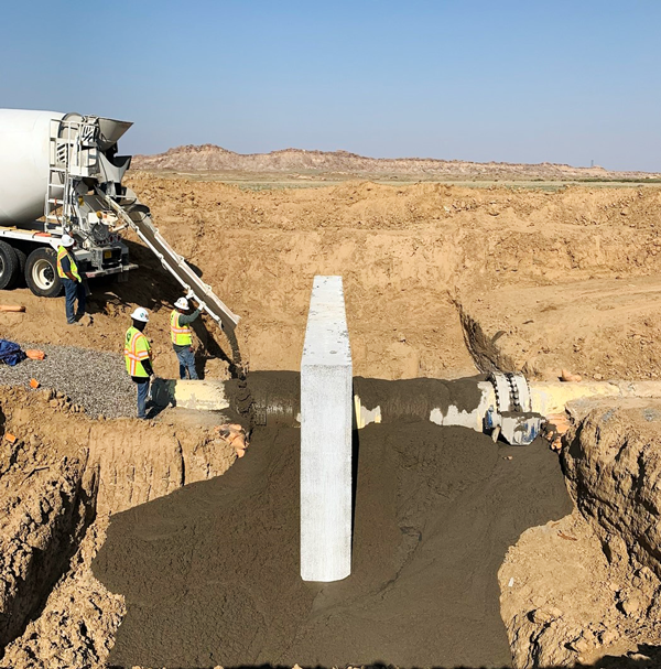Two construction workers pouring concrete to backfill around a pieline collar in the New Mexico desert.