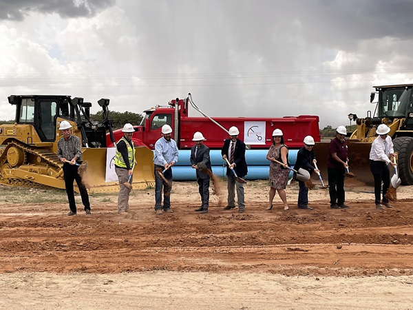 Outdoor view of nine professionals in hardhats turning dirt with shovels.  Several pieces of heavy equipment are parked in the background. 