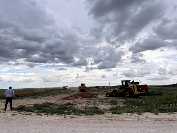 Outdoor view of dirt terrain and brush with a few heavy equipment vehicles. A construction manager is standing at left. 