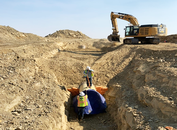 Two construction workers installing a 42-inch steel pipe in a trench in the New Mexico desert with an excavator in the background.