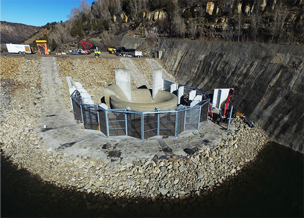 The fish screen is installed around the spillway and there are construction vehicles in the background.