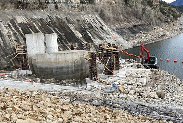 A red excavator works on the foundation of the fish screen with rocks and boulders all around and rip rap in place around the spillway.
